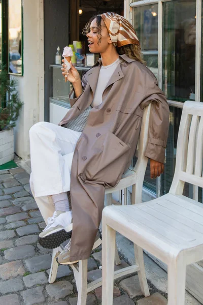 Здивована афроамериканська жінка в хедкарфі і стильна траншея пальто тримає морозиво конус і сидить на стільці зовні — стокове фото