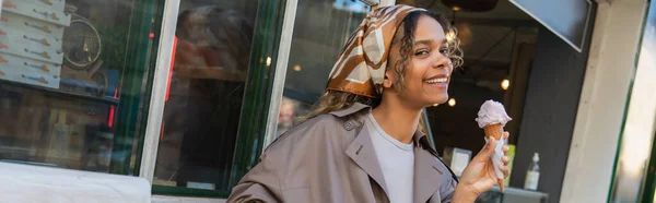 Cheerful african american woman in headscarf and stylish trench coat holding ice cream in prague, banner - foto de stock