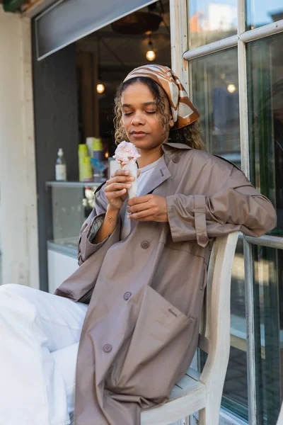 Young african american woman in headscarf looking at ice cream cone and sitting on chair in prague - foto de stock