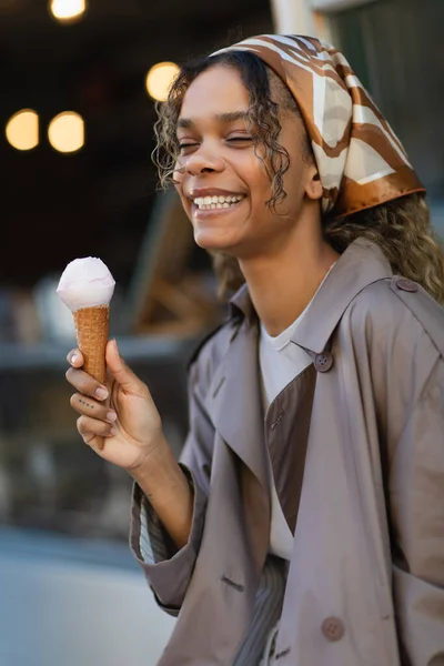 Smiling african american woman in headscarf holding ice cream cone and sitting on chair in prague — Stockfoto