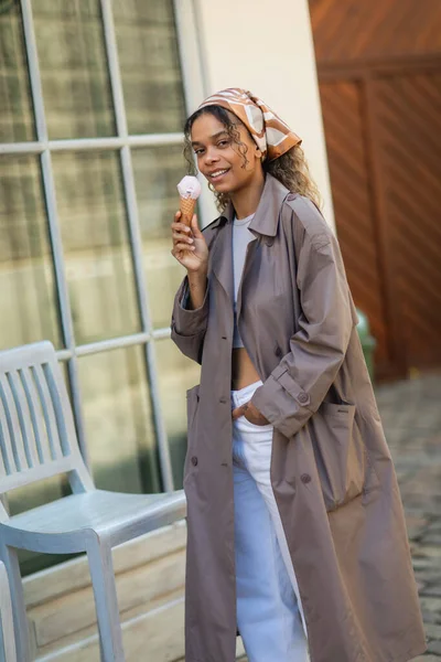 Cheerful african american woman in headscarf and stylish trench coat holding ice cream cone on street in prague — Stockfoto