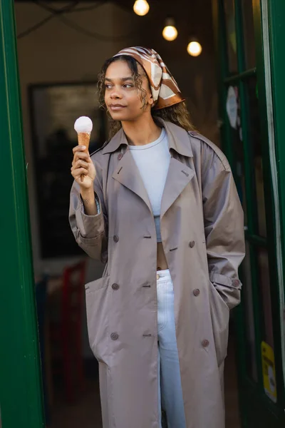Young african american woman in headscarf and stylish trench coat holding ice cream cone on street in prague — Fotografia de Stock