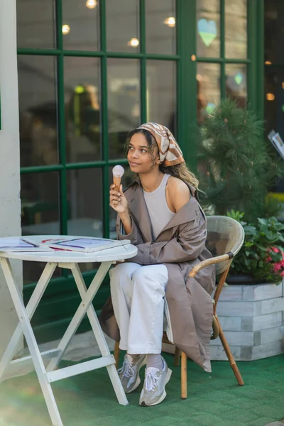 Татуированная африканская американка в платке и стильном плаще держит мороженое с конусом и сидит на террасе кафе — стоковое фото
