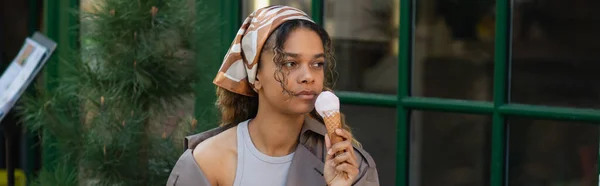 Young african american woman in headscarf holding ice cream cone and sitting in cafe terrace, banner — Stockfoto