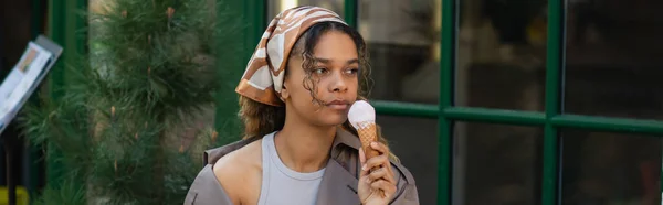 Young african american woman in headscarf holding ice cream cone and sitting in cafe terrace, banner — Stock Photo