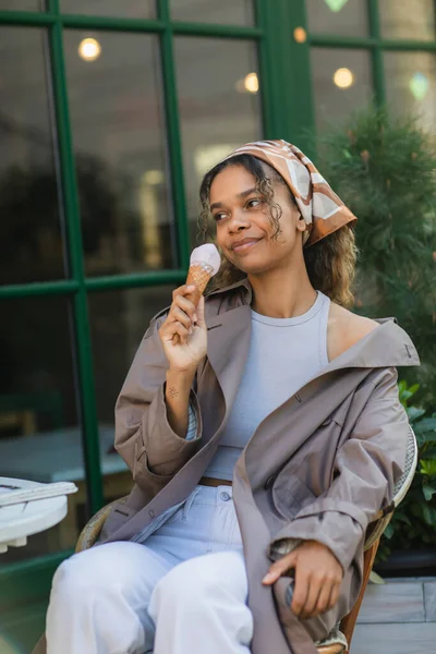 Smiling african american woman in headscarf and stylish trench coat holding ice cream cone and sitting in cafe terrace — Foto stock