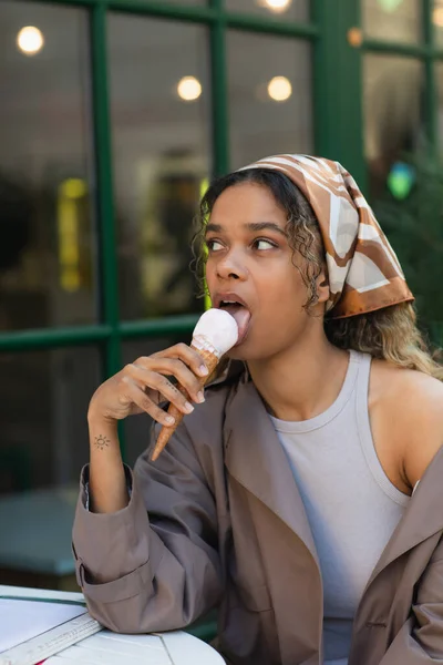 Tattooed african american woman in headscarf and stylish trench coat eating ice cream and sitting in cafe terrace — Fotografia de Stock