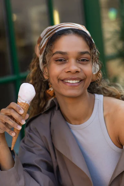 Happy african american woman in headscarf holding ice cream cone and sitting in cafe terrace — Foto stock