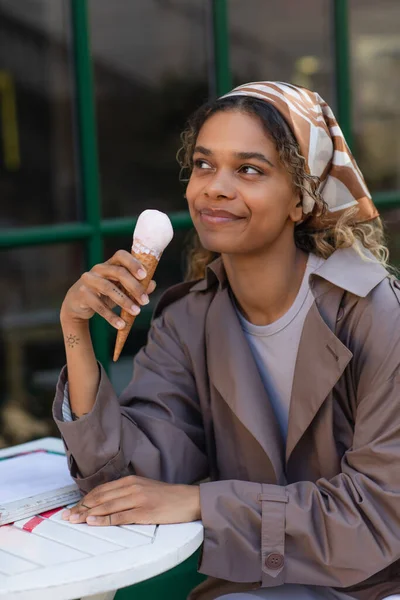 Smiling african american woman in stylish trench coat holding ice cream cone and sitting in cafe terrace - foto de stock