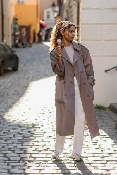 Full length of young african american woman in stylish trench coat and headscarf holding ice cream cone on street in prague — Stockfoto