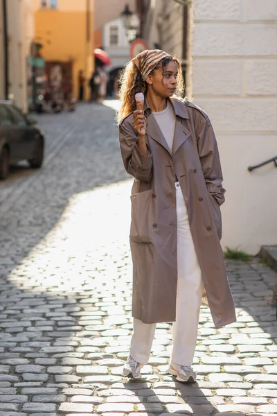 Full length of young african american woman in stylish trench coat and headscarf holding ice cream cone on street in prague — Stock Photo