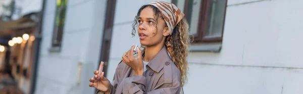 Young african american woman in stylish trench coat and headscarf applying lip gloss on street in prague, banner — Stockfoto