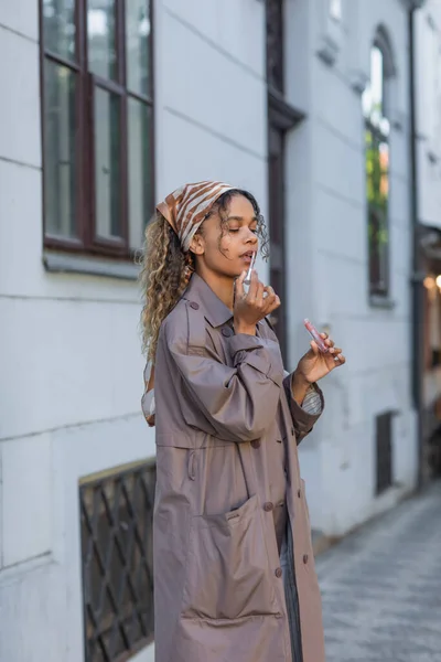 Curly african american woman in stylish trench coat and headscarf applying lip gloss on street in prague - foto de stock