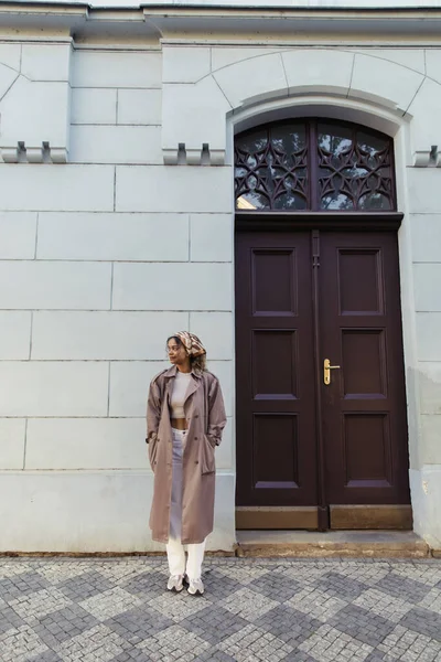 Full length of african american woman in trendy outfit and headscarf standing with hands in pockets near building in prague — Fotografia de Stock