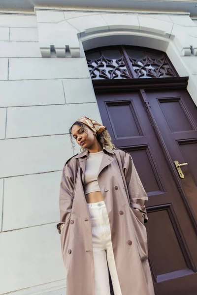 Low angle view of african american woman in trendy outfit and headscarf standing with hands in pockets near building in prague — Fotografia de Stock