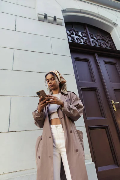African american woman in trendy outfit and headscarf using smartphone near building in prague — Stockfoto