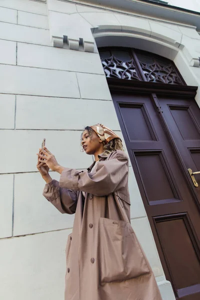 African american woman in trendy outfit and headscarf taking photo on smartphone near building in prague — Fotografia de Stock