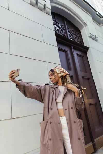African american woman in trendy outfit and headscarf taking selfie on smartphone near building in prague — Stockfoto