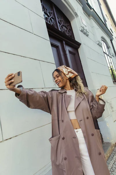 Happy african american woman in trendy outfit taking selfie on smartphone near building in prague — Fotografia de Stock