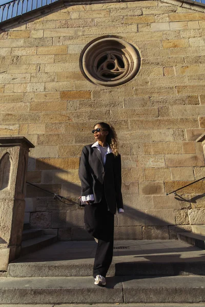 Full length of curly african american woman in sunglasses and oversize suit standing near ancient wall on street in prague — Stock Photo