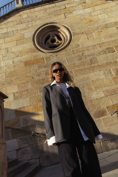 Low angle view of stylish african american woman in sunglasses and oversize suit standing near ancient wall on street in prague — Stock Photo