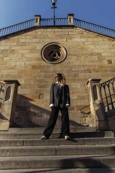Full length of stylish african american woman in sunglasses and oversize suit standing near ancient wall on street in prague — Stock Photo