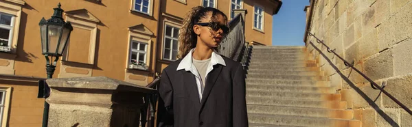 Stylish african american woman in sunglasses and oversize suit standing near stairs on street in prague, banner — Stockfoto
