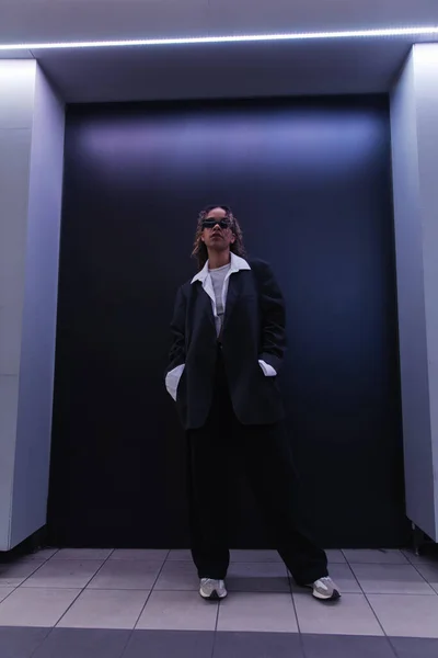 Stylish african american woman in sunglasses and oversize suit posing with hands in pockets near dark blue wall — Foto stock