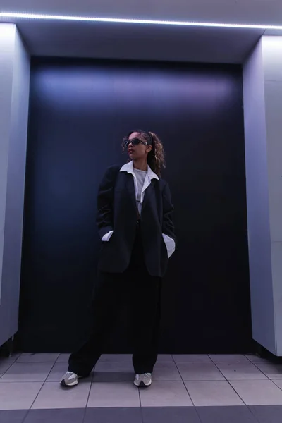 Full length of african american woman in sunglasses and oversize suit posing with hands in pockets near dark blue wall — Fotografia de Stock