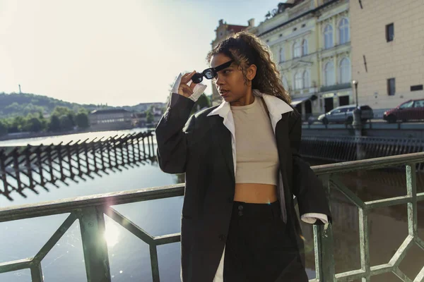 Stylish african american woman in oversize suit wearing sunglasses and standing near river in prague — Photo de stock