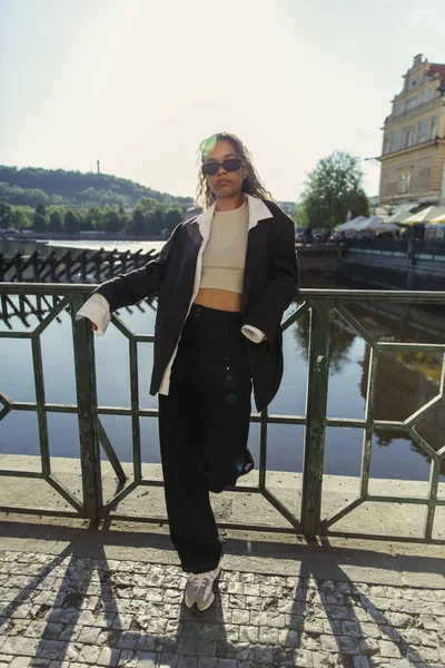 Full length of stylish african american woman in oversize suit and sunglasses standing near river in prague — Stock Photo