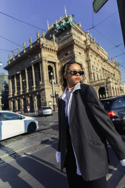 Кудрявая африканская американка в огромных пиджаках и солнцезащитных очках стоит рядом с национальным театром в Праге — стоковое фото