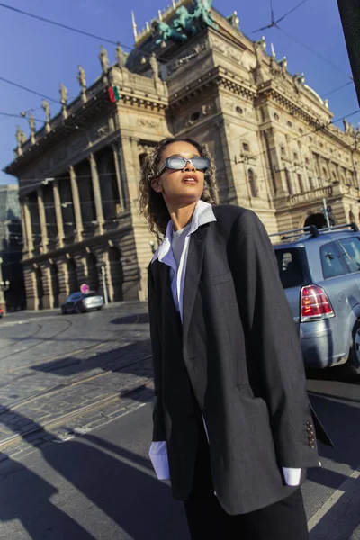 Стильная африканская американка в огромном пиджаке и солнцезащитных очках, стоящая рядом с национальным театром в Праге — стоковое фото