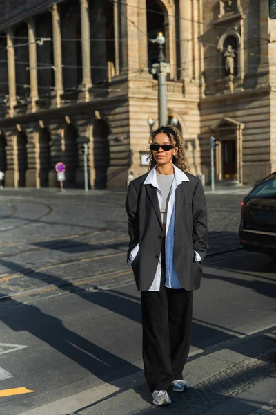 Curly african american woman in oversized blazer and sunglasses walking with hands in pockets near building in prague - foto de stock