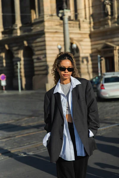 Young african american woman in stylish blazer and sunglasses walking with hands in pockets near building in prague — Stock Photo