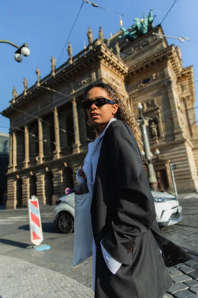 Low angle view of young african american woman in stylish blazer and sunglasses near national theatre in prague — Fotografia de Stock