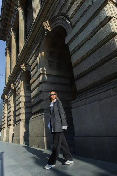 Young african american woman in sunglasses and oversize suit walking near building on urban street in prague — Stockfoto