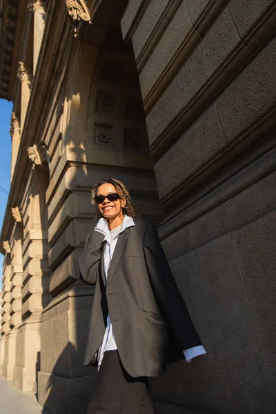 Sunshine on face of happy african american woman in sunglasses and oversize suit walking on urban street in prague — Foto stock