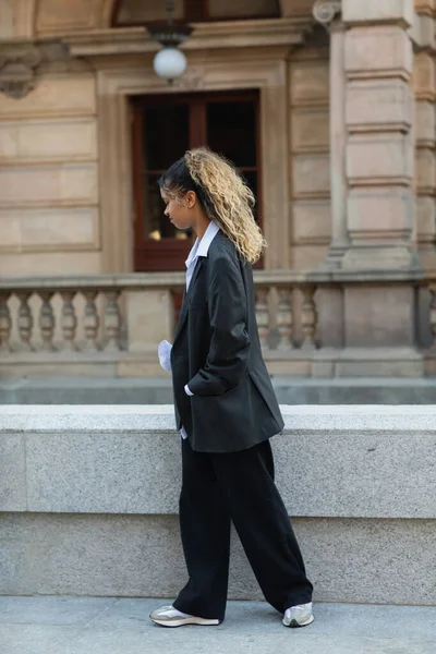 Side view of displeased african american woman in oversize suit walking with hand in pocket on urban street in prague — Stockfoto