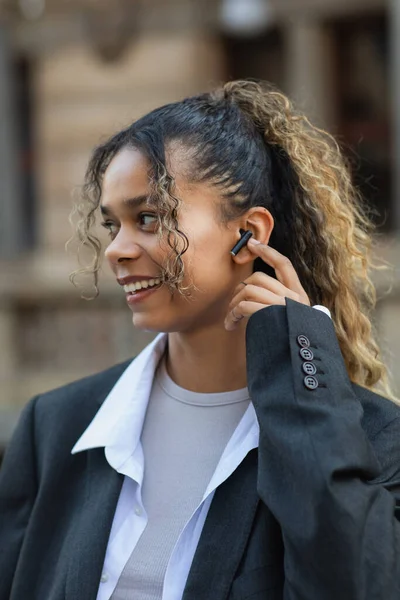 Happy african american woman in oversize suit adjusting wireless earphone on urban street of prague — Stock Photo