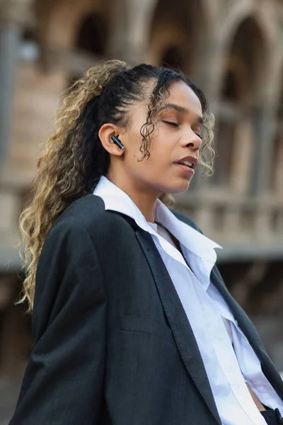 African american woman with closed eyes listening music in wireless earphone on urban street of prague — Foto stock