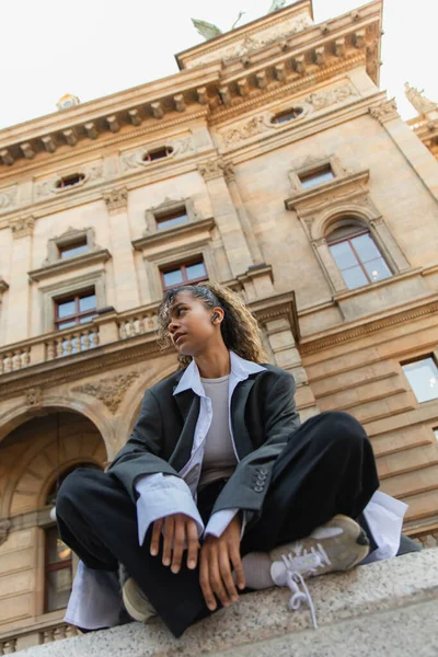 Bottom view of african american woman in oversize suit listening music in wireless earphone near national theatre in prague — Stockfoto