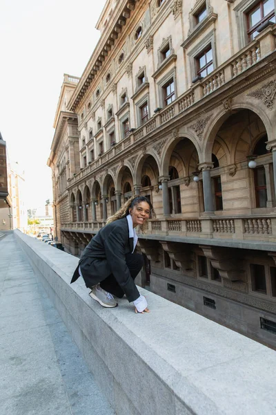 Positive african american woman in oversize suit listening music in wireless earphone while sitting near building in prague — Stockfoto