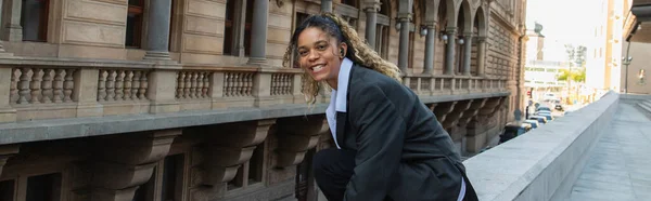 Positive african american woman in oversize suit listening music in wireless earphone while sitting near building in prague, banner - foto de stock