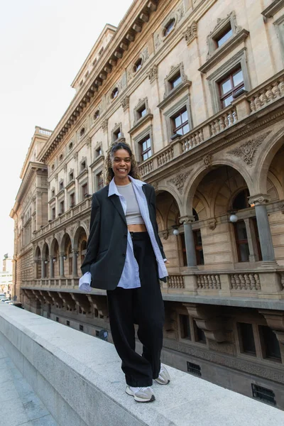 Full length of positive african american woman in oversize suit walking near building of national theater in prague - foto de stock