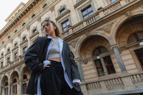 Low angle view of african american woman in wireless earphone posing on urban street near building of national theater in prague — Foto stock