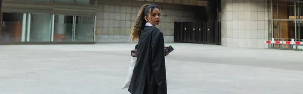 African american woman in oversize suit listening music in wireless earphone and walking on urban street in prague, banner — Stockfoto