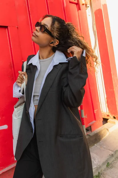 African american woman in oversize suit and sunglasses listening music and standing with reusable bag on urban street in prague — Fotografia de Stock