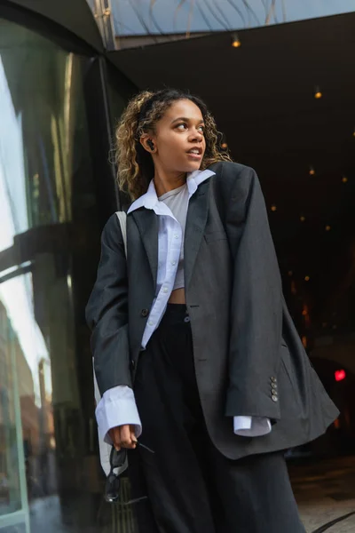 African american woman in oversize blazer holding sunglasses while listening music on urban street in prague — Photo de stock