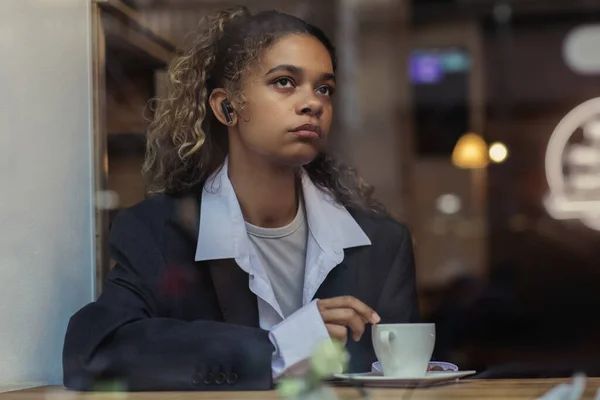 Sad african american woman in wireless earphone listening music near cup of coffee in cafe — Foto stock